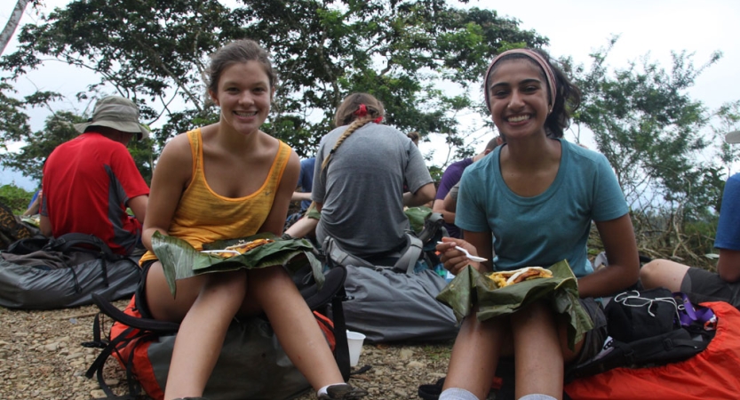 Two people sit on their backpacks while eating a snack. They smile for the photo. 